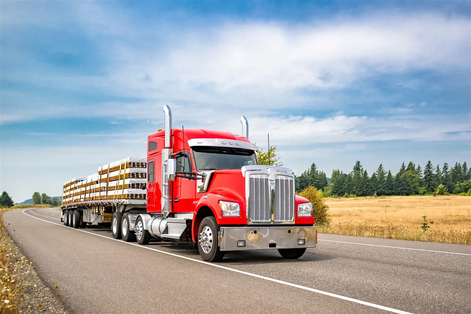 Red semi-truck with a flatbed trailer loaded with lumber on a two-lane highway