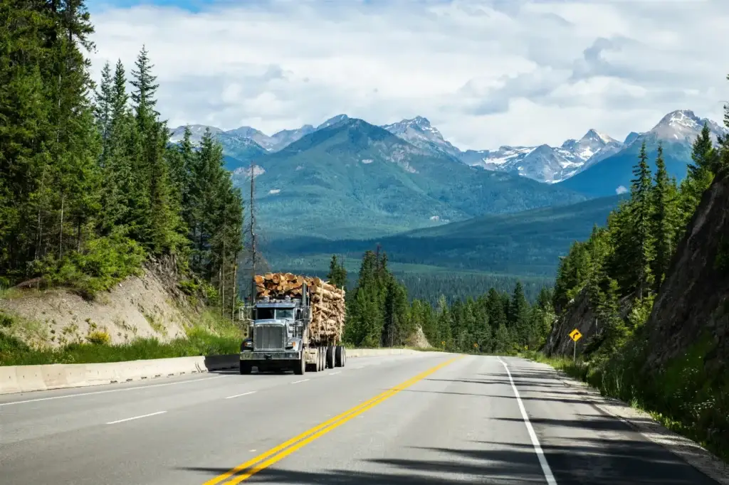 Log truck on a mountain highway with evergreen forest and snowy peaks